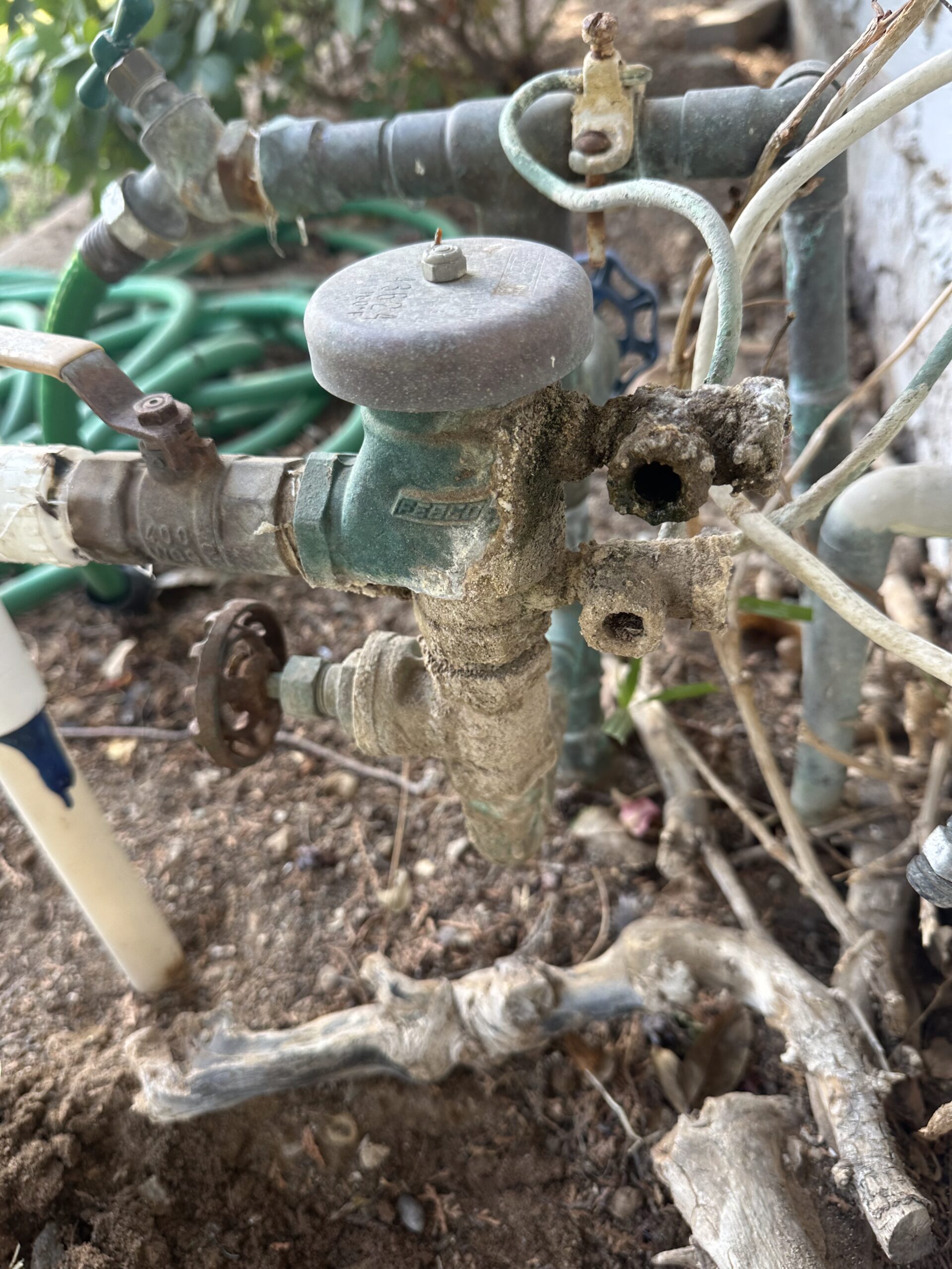 Leaking and corroded main water supply valve with visible damage, surrounded by plumbing pipes and dirt, highlighting plumbing issues in Phoenix, AZ.