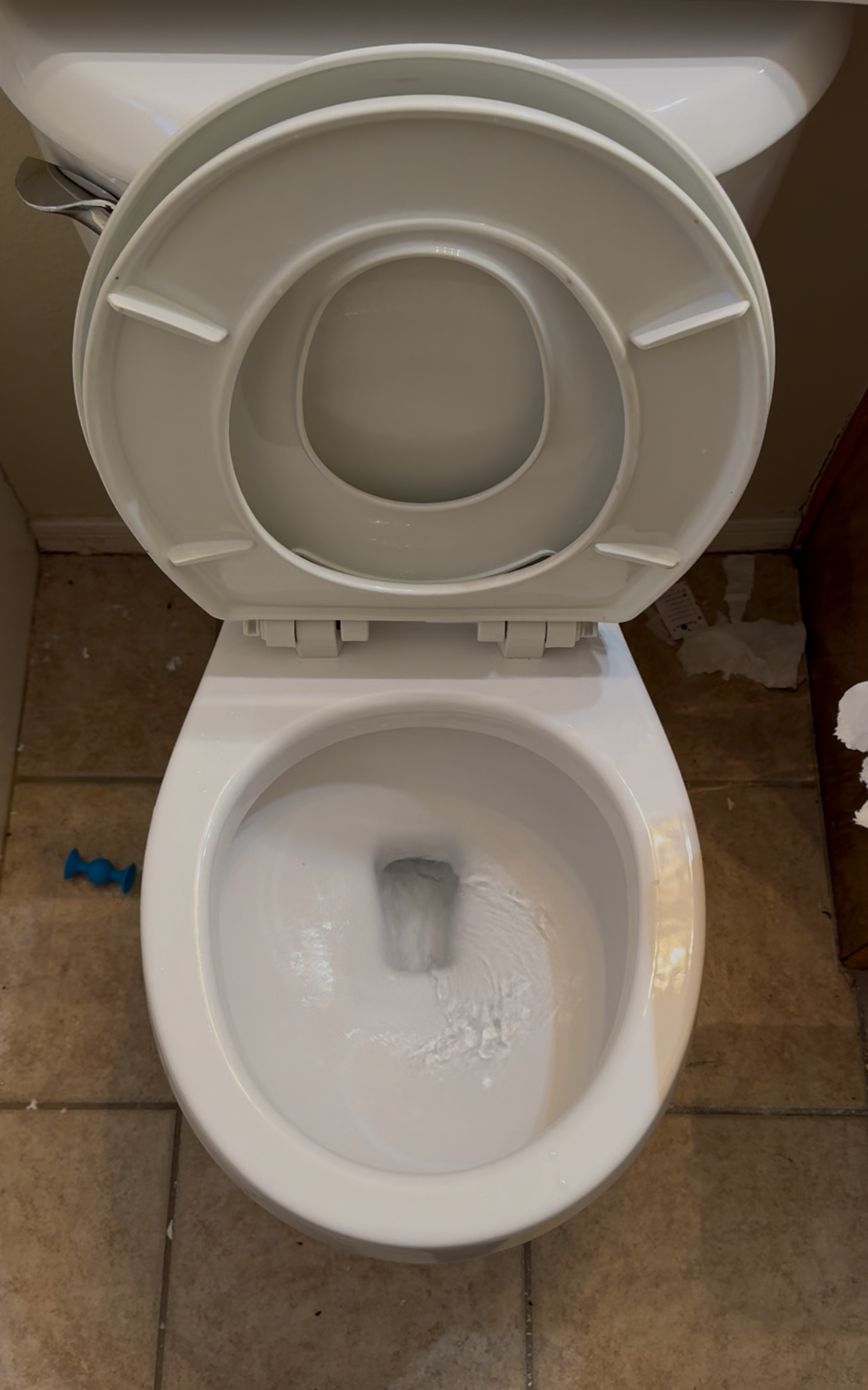 Toilet viewed from above, showing clean water in the bowl and an open lid, with a blue toy visible on the tiled floor, relevant to plumbing services and toilet replacement discussions.
