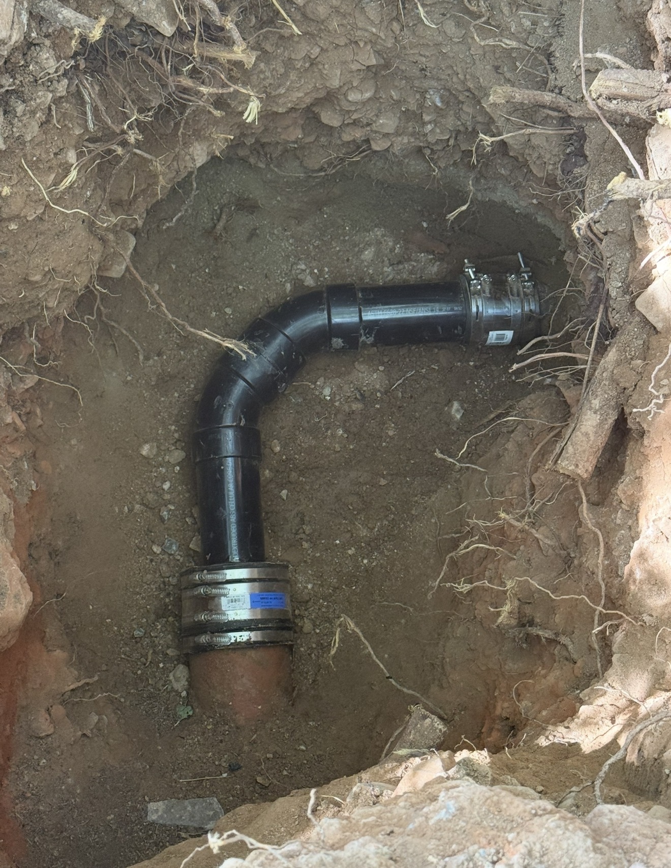 Sewer pipe installation in a trench, featuring black plastic piping and metal couplings, surrounded by soil and roots, relevant to sewer repair services in West Valleys, AZ.