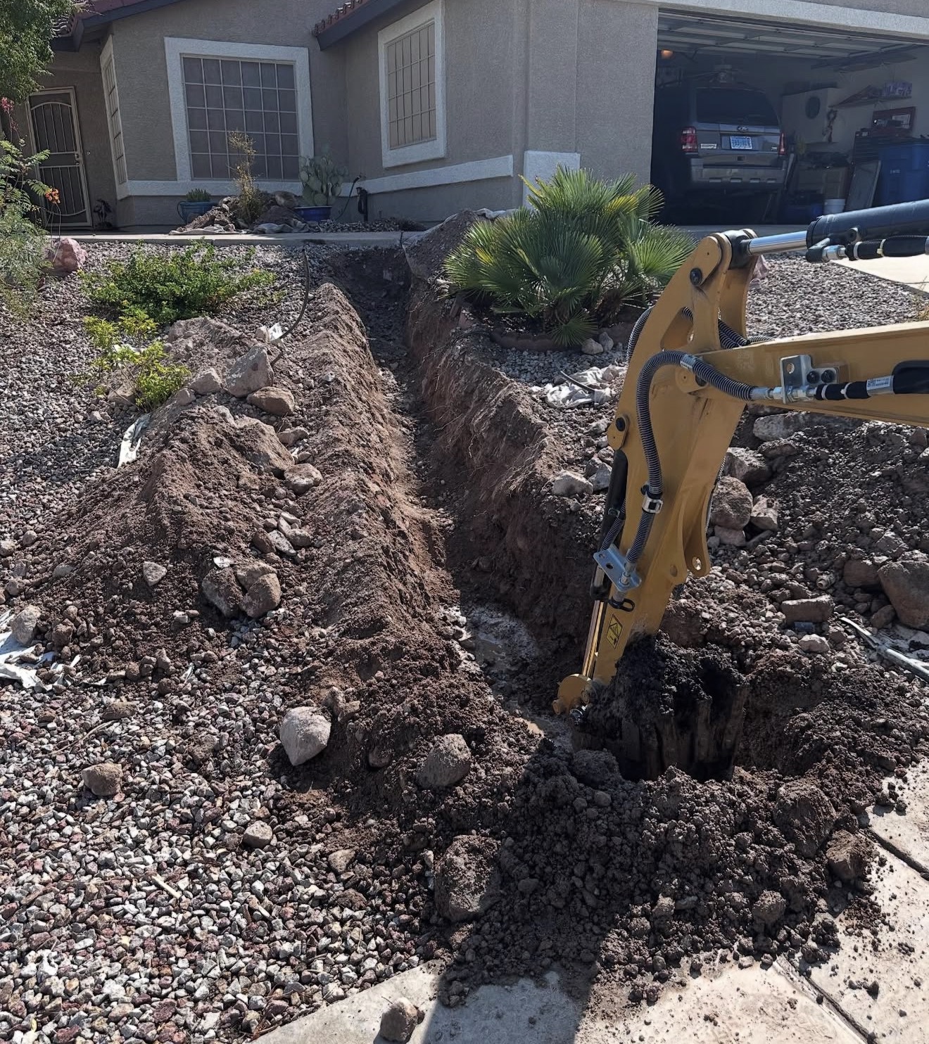 Excavation site in front of a house with a trench for plumbing work, showcasing a digging machine and disturbed soil, relevant to plumbing services in Surprise, AZ.