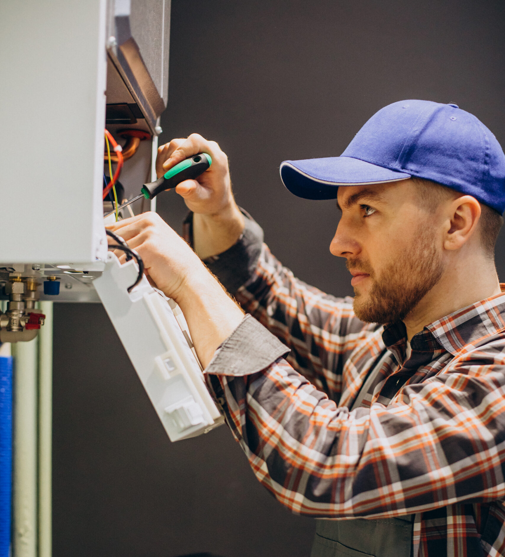 service-man-adjusting-house-heating-system-scaled-e1741996498521.jpg Technician repairing a water heating system, focusing on internal components with a screwdriver, wearing a blue cap and plaid shirt, in a professional plumbing service setting.